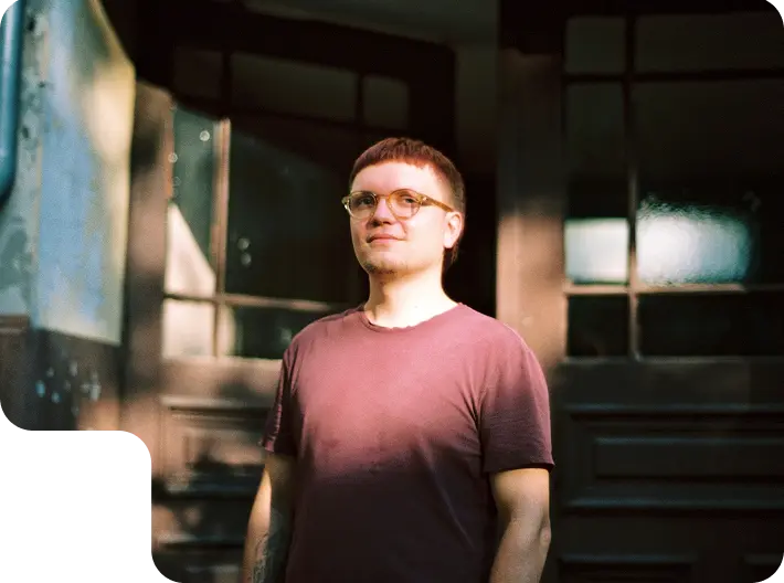A portrait photo of Simon Schötz in a wine-red shirt and round glasses. He is standing in front of big doors with glass windows, one of which is partially open.